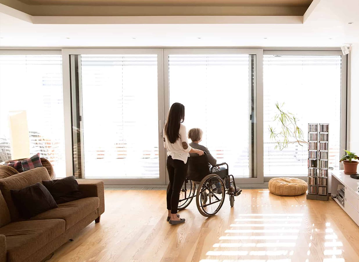 Ein Frau steht mit einer anderen Frau im Rollstuhl am Fenster in einem geräumigen Wohnzimmer